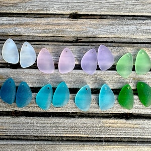 Puede incluir: Una variedad de colgantes de vidrio marino en varios colores, incluyendo azul claro, lavanda, turquesa y verde. Cada pieza tiene una forma orgánica única y un pequeño agujero para ensartar. Los colgantes están dispuestos sobre una superficie de madera envejecida.