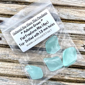 May include: A clear plastic bag containing four sea glass pendants. The pendants are a light teal colour and have a flat, freeform shape. The bag has a white label with the text "Cultured Sea Glass Flat Pendants" and "Autumn Green". Each pendant is drilled with 1.5 mm holes.