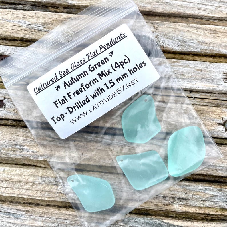 May include: A clear plastic bag containing four sea glass pendants. The pendants are a light teal colour and have a flat, freeform shape. The bag has a white label with the text "Cultured Sea Glass Flat Pendants" and "Autumn Green". Each pendant is drilled with 1.5 mm holes.