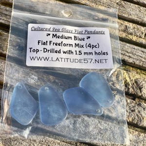 May include: Four medium blue, flat, freeform sea glass pendants in a clear plastic bag. The pendants are top-drilled with 1.5 mm holes. The bag has a label that reads "Cultured Sea Glass Flat Pendants" and "Medium Blue."
