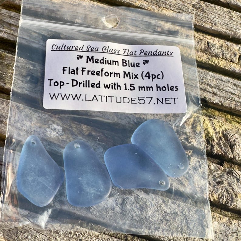 May include: Four medium blue, flat, freeform sea glass pendants in a clear plastic bag. The pendants are top-drilled with 1.5 mm holes. The bag has a label that reads "Cultured Sea Glass Flat Pendants" and "Medium Blue."