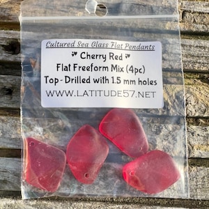 May include: A transparent bag holding four cherry red sea glass pendants. The pendants are flat and freeform, with 1.5 mm holes drilled at the top. The bag is labelled "Cultured Sea Glass Flat Pendants" and "Cherry Red".