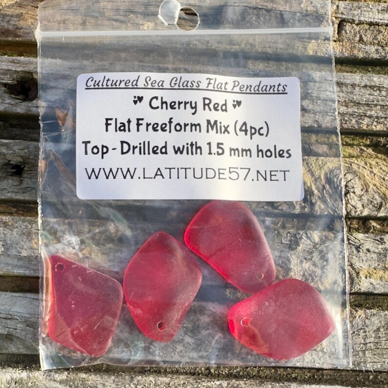May include: A transparent bag holding four cherry red sea glass pendants. The pendants are flat and freeform, with 1.5 mm holes drilled at the top. The bag is labelled "Cultured Sea Glass Flat Pendants" and "Cherry Red".