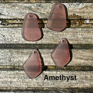 May include: Four amethyst-coloured sea glass pendants with a hole at the top for stringing. The pendants are various organic shapes and sit on a weathered wooden surface. The word "Amethyst" is printed in black.
