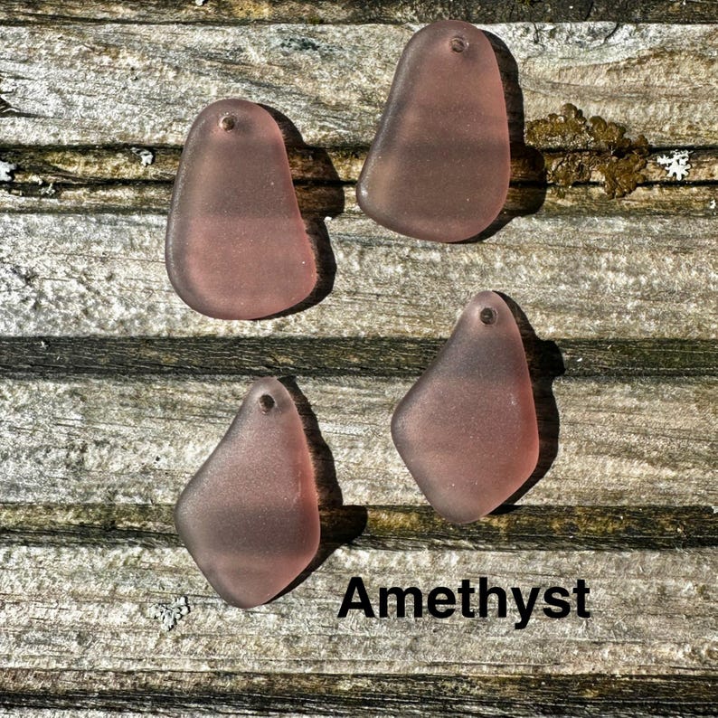 May include: Four amethyst-coloured sea glass pendants with a hole at the top for stringing. The pendants are various organic shapes and sit on a weathered wooden surface. The word "Amethyst" is printed in black.