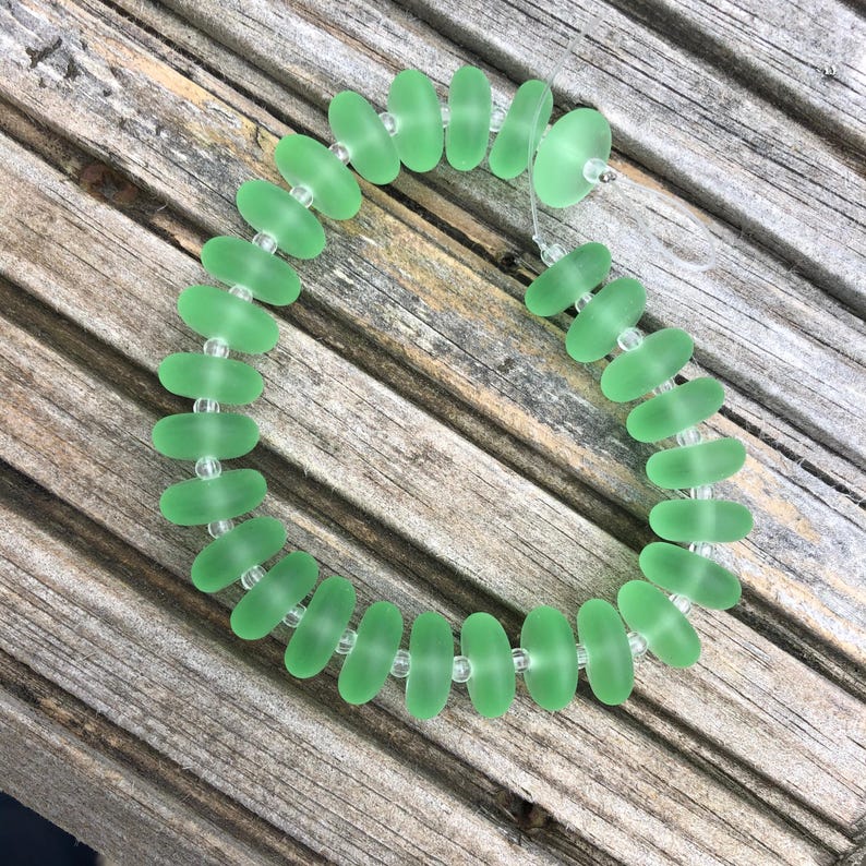 May include: A handmade bracelet featuring oval-shaped, seafoam green beads. The beads are strung together with clear, small beads. The bracelet is displayed on a weathered, wooden surface.