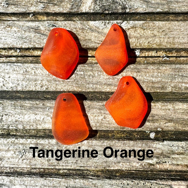 May include: Four tangerine orange sea glass pendants with a hole drilled at the top, arranged on a weathered wooden surface. The text "Tangerine Orange" is displayed below the pendants.