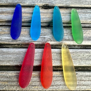 May include: Seven teardrop-shaped sea glass pendants in various colours, including blue, green, red, and yellow. Each has a small hole for stringing. The pendants are displayed on a weathered wooden surface.