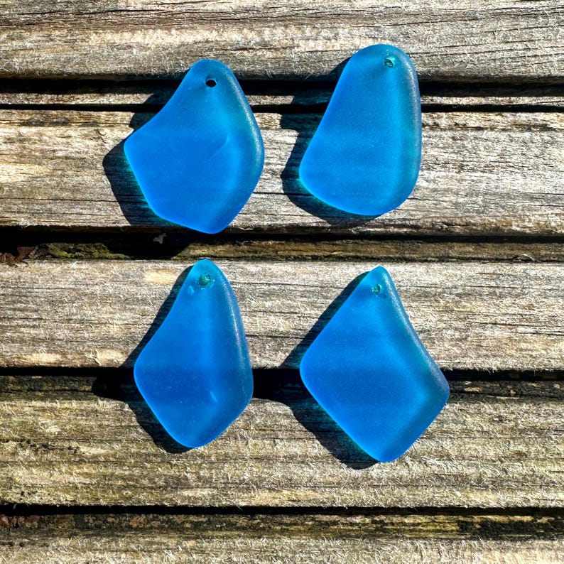 May include: Four pieces of sea glass in a vibrant blue hue, each with a unique, organic shape and a small hole at the top. The sea glass is displayed on a weathered wooden surface, creating a contrast of textures and colours.