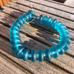 May include: A turquoise beaded bracelet featuring a combination of round and flattened beads. The beads are strung on a clear cord, creating a delicate piece of jewellery. The bracelet is displayed on a weathered wooden surface.