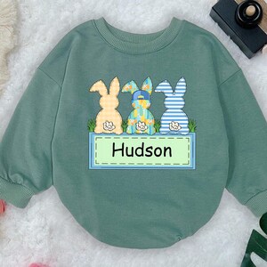 May include: Sage green baby sweatshirt with three cartoon bunny rabbits and the name "Hudson" on a rectangular sign. The bunnies have different patterns and colors, with one wearing a hat. The sweatshirt has long sleeves.
