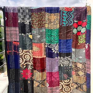 May include: A patchwork quilt with a variety of patterned squares in shades of blue, red, green, and brown. The quilt is hanging outside, with a black border and white stitching throughout. The quilt is made of various fabrics.