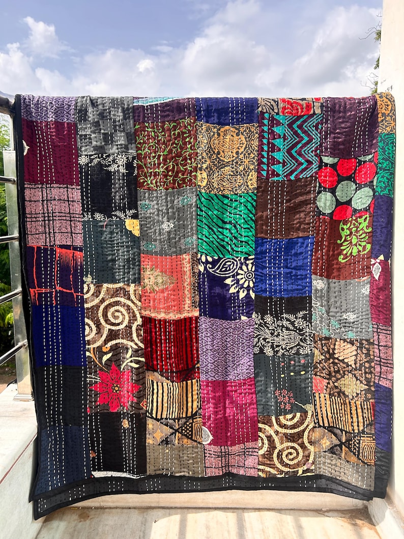 May include: A patchwork quilt with a variety of patterned squares in shades of blue, red, green, and brown. The quilt is hanging outside, with a black border and white stitching throughout. The quilt is made of various fabrics.