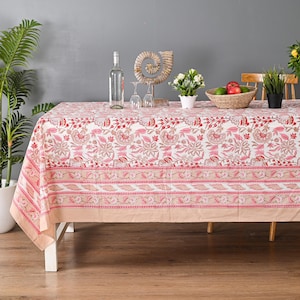 Könnte beinhalten: Eine rechteckige Tischdecke mit einem Blumenmuster in Rosa- und Rottönen, auf einem weißen Tisch. Die Tischdecke hat einen Rand mit einem Blattdesign. Eine Flasche, Gläser und Dekorationsgegenstände stehen auf dem Tisch.