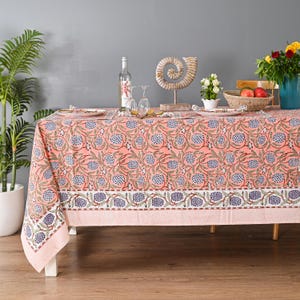 Könnte beinhalten: Eine korallenfarbene Tischdecke mit floralem Muster, auf einem Holztisch. Die Tischdecke hat ein sich wiederholendes Design aus blauen und grünen floralen Elementen. Ein weißer Rand mit Streifenmuster ist sichtbar. Der Tisch ist mit Tellern, Gläsern und einer dekorativen Muschel gedeckt.