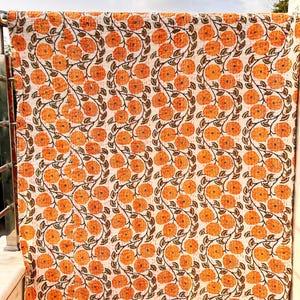 Puede incluir: Un edredón con estampado floral blanco y naranja. El edredón presenta un patrón repetitivo de flores naranjas con tallos y hojas marrones. El edredón está colgado de una barandilla de metal, con un cielo azul de fondo.