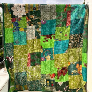 May include: A patchwork quilt with a vibrant mix of green hues, including lime, emerald, and forest green. The quilt features a variety of floral and geometric patterns, creating a textured and visually appealing design. The quilt is displayed outdoors, with a clear sky in the background.
