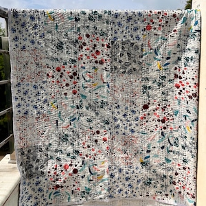 Puede incluir: Un edredón blanco con un estampado floral en tonos rojos, azules, verdes y grises. El edredón está colgado al aire libre, con costuras visibles. El diseño combina elementos botánicos y abstractos.