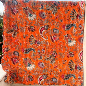 Könnte beinhalten: Eine orangefarbene Steppdecke mit einem detaillierten Blumen- und Paisley-Muster in Rot-, Blau- und Weißtönen. Die Steppdecke hängt im Freien und zeigt ihr aufwendiges Design und ihre Nähte. Das Gesamtthema ist böhmisch und dekorativ.