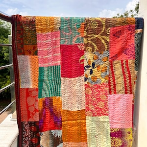 Könnte beinhalten: Ein Patchwork-Quilt mit einer Vielzahl von bunten Stoffquadraten. Der Quilt zeigt eine Mischung aus floralen, gemusterten und unifarbenen Stoffen in Rot-, Orange-, Grün- und Rosatönen. Die Kanten sind mit einem dunkelroten Rand versehen.
