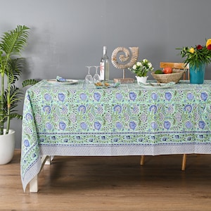 May include: A rectangular tablecloth with a floral pattern in shades of blue and green. The tablecloth is set on a wooden table with a white base. The table is set with plates, glasses, and napkins.