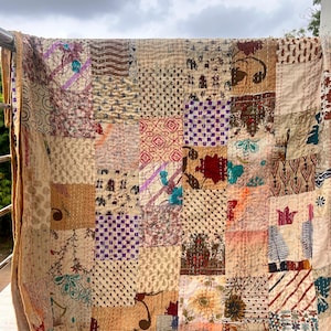 Könnte beinhalten: Ein Patchwork-Quilt mit einer Vielzahl von gemusterten Quadraten in Beige-, Braun- und gedämpften Farben. Der Quilt ist über ein Geländer drapiert und zeigt sein aufwendiges Design und seine Nähte. Die Ränder des Quilts sind einfarbig beige.