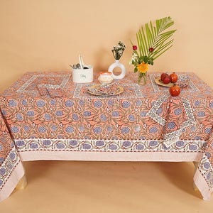 Könnte beinhalten: Ein rechteckiges, korallenfarbenes Tischtuch mit floralem Muster, gedeckt für eine Mahlzeit. Das Muster umfasst blaue und weiße Blumendesigns. Der Tisch ist mit passenden Servietten, Tellern und einer Schüssel gedeckt. Eine Vase mit Blumen und Früchten vervollständigt die Szene.