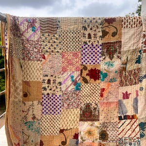 Puede incluir: Un edredón de retazos con una variedad de cuadrados estampados en beige, marrón y colores apagados. El edredón está colgado de una barandilla, mostrando su intrincado diseño y costuras. Los bordes del edredón son de color beige liso.