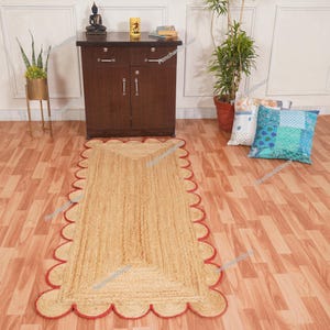Könnte beinhalten: Ein rechteckiger Jute-Teppich mit einem roten, wellenförmigen Rand. Der Teppich hat eine natürliche beige Farbe und liegt auf einem Holzboden. Der Teppich ist ein dekoratives Element für ein Zuhause.