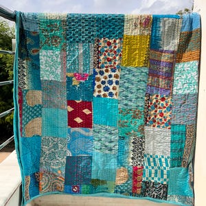 May include: A patchwork quilt with a variety of blue, teal, and turquoise fabrics. The quilt is made of squares and rectangles of different patterns and textures, with a solid turquoise border. The quilt is hanging on a metal railing.