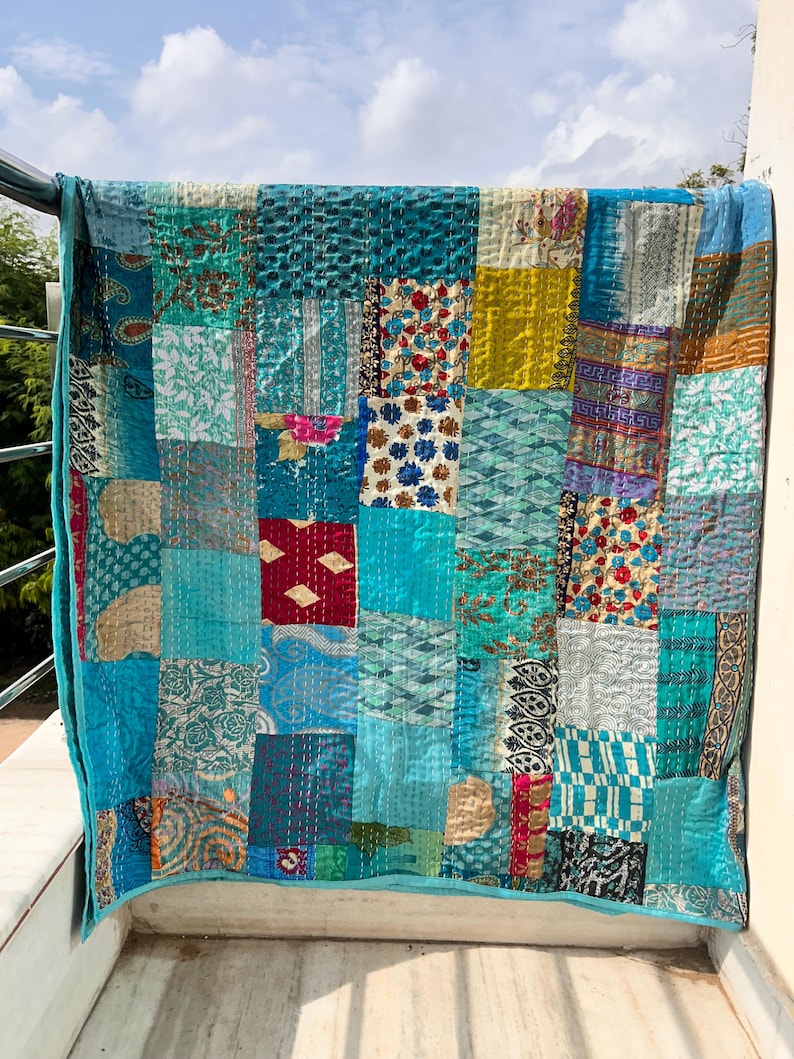 May include: A patchwork quilt with a variety of blue, teal, and turquoise fabrics. The quilt is made of squares and rectangles of different patterns and textures, with a solid turquoise border. The quilt is hanging on a metal railing.