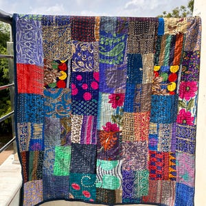 May include: A patchwork quilt with a variety of fabric patterns and colors, including blue, purple, red, and gold. The quilt is hanging on a railing, with a blue sky in the background. The quilt is made of squares of fabric sewn together.