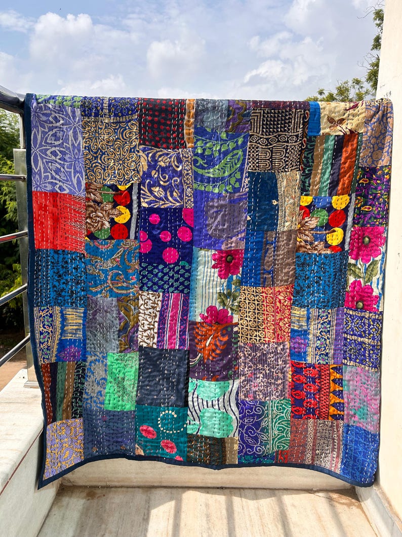 May include: A patchwork quilt with a variety of fabric patterns and colors, including blue, purple, red, and gold. The quilt is hanging on a railing, with a blue sky in the background. The quilt is made of squares of fabric sewn together.