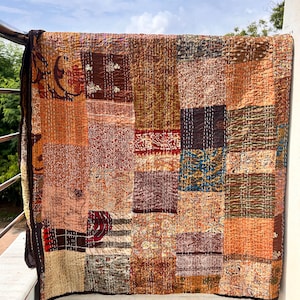 May include: A patchwork quilt with a variety of warm colors, including shades of brown, orange, and red. The quilt is made of rectangular fabric pieces with different patterns and textures. The quilt is hanging on a railing.