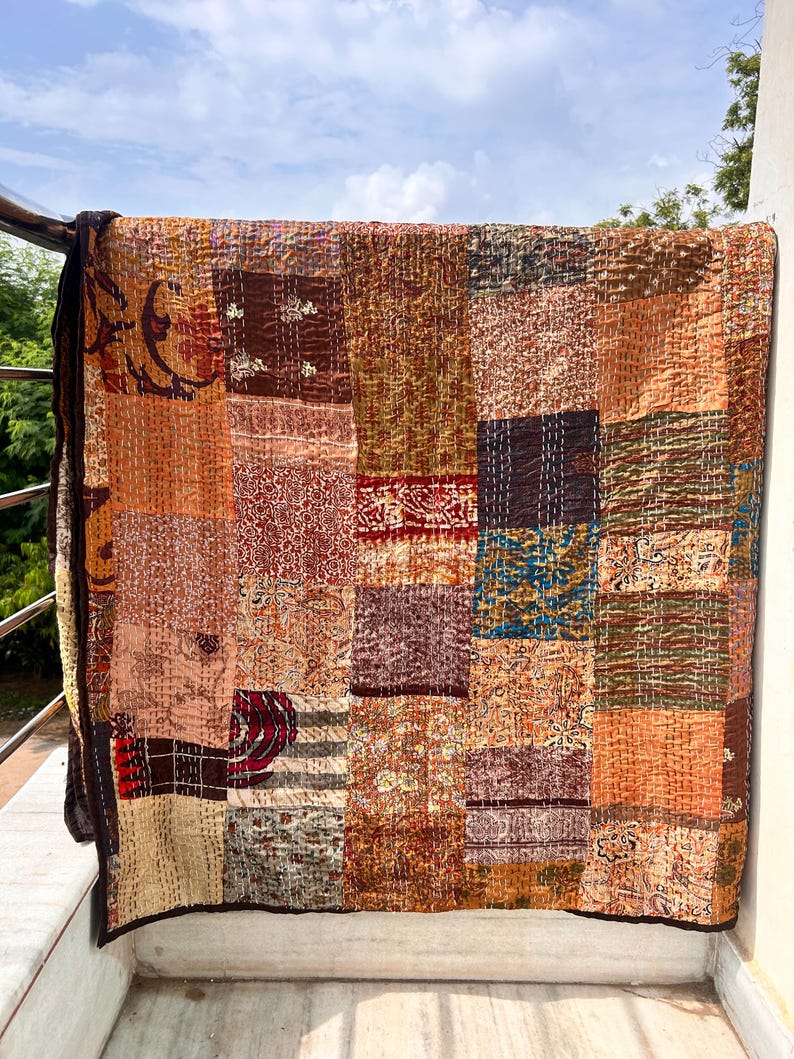 May include: A patchwork quilt with a variety of warm colors, including shades of brown, orange, and red. The quilt is made of rectangular fabric pieces with different patterns and textures. The quilt is hanging on a railing.