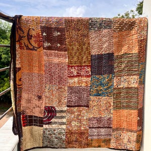 Könnte beinhalten: Ein Patchwork-Quilt in warmen Farben wie Braun, Orange und Rot. Der Quilt besteht aus rechteckigen Stoffstücken mit verschiedenen Mustern und Texturen. Er hängt an einem Metallgeländer und zeigt sein aufwendiges Design.