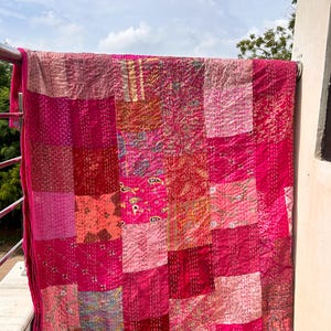 May include: A vibrant, patchwork quilt in shades of pink and red, hanging outdoors. The quilt features a variety of patterned fabrics stitched together, creating a textured and colorful design. The edges are finished with a solid pink border.
