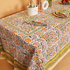 Puede incluir: Un mantel blanco con un vibrante estampado floral en tonos rojos, amarillos y verdes. La mesa está puesta con manteles individuales y servilletas a juego, creando una estética de comedor cohesiva. También se ven un portacubiertos y jarrones decorativos.