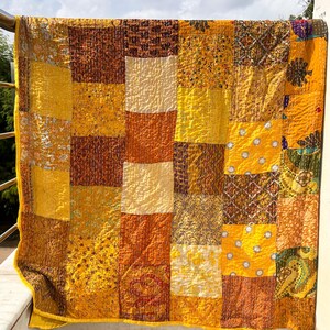 May include: A patchwork quilt with a vibrant mix of yellow, orange, and brown squares. The quilt is made of various patterned fabrics, creating a textured and visually appealing design. The quilt is hanging on a railing, with a sky background.