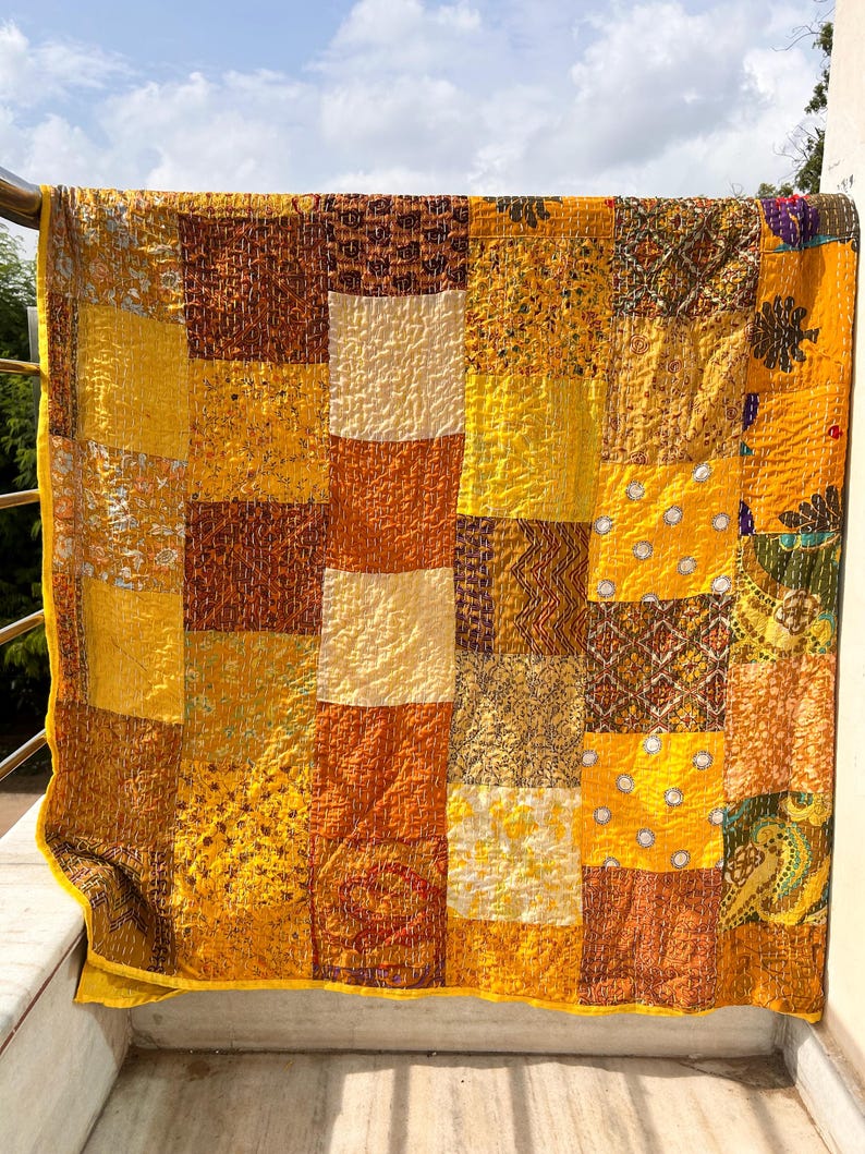 May include: A patchwork quilt with a vibrant mix of yellow, orange, and brown squares. The quilt is made of various patterned fabrics, creating a textured and visually appealing design. The quilt is hanging on a railing, with a sky background.