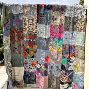May include: A patchwork quilt with a variety of fabric patterns and colors, including gray, blue, pink, and floral designs. The quilt is hanging outdoors, showcasing its stitched construction and unique design. The quilt is a decorative home textile.