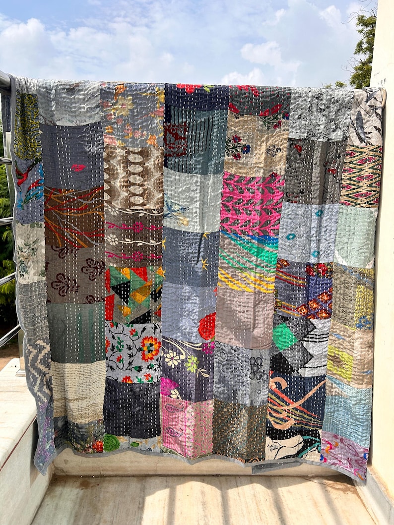 May include: A patchwork quilt with a variety of fabric patterns and colors, including gray, blue, pink, and floral designs. The quilt is hanging outdoors, showcasing its stitched construction and unique design. The quilt is a decorative home textile.