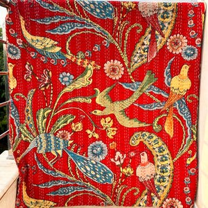 May include: A red textile with a vibrant floral and bird pattern. The design features birds, flowers, and foliage in shades of yellow, blue, and green. The fabric is stitched with a repeating pattern.