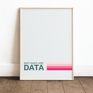 May include: Framed art print with the words "MISTAKES ARE DATA" in teal, with a pink and magenta color block design. The print is in a gold frame and is displayed against a white wall and wooden floor.