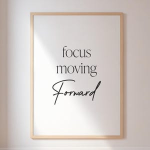 Pode incluir: Uma impressão emoldurada com as palavras "focus moving Forward" em preto, em fontes de script e sem serifa. A impressão tem um fundo branco e uma moldura de cor de madeira clara. A estética geral é minimalista e motivacional.