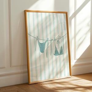 May include: A framed print featuring a teal bikini line drawing hanging on a clothesline against a striped background. The artwork is in a wooden frame and leans against a white wall, with sunlight casting shadows on the floor.