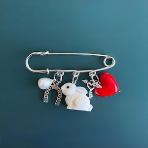 May include: A silver safety pin charm with dangling charms. Charms include a white rabbit, a red heart, a horseshoe with "GOOD LUCK" text, and a key. The pin is displayed against a teal background.