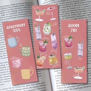 May include: Pink and peach-colored cocktail recipe cards with various drink illustrations and names. The cards are set against the backdrop of an open book, creating a vintage aesthetic.