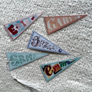 May include: Five colorful pennant flags with names printed on them. The flags are in shades of pink, peach, blue, and teal. Each flag has a different name in a unique font. One flag has a tassel attached.
