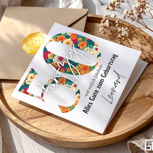 Può includere: Un biglietto di compleanno con il numero 50 in un design floreale, con la parola "funtzig" in corsivo. Il biglietto è su un vassoio di legno con una busta e un sigillo dorato. Il testo recita "Alles Gute zum Geburtstag Leonard".